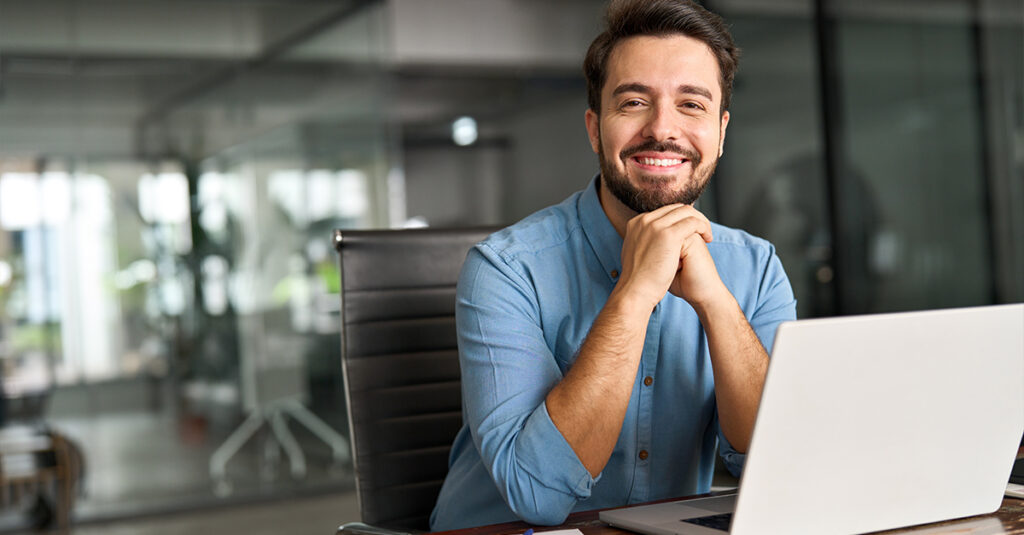 Homme professionnel devant un ordinateur, souriant à la caméra, illustrant le sujet du blog sur Substack comme ressource d’apprentissage des langues